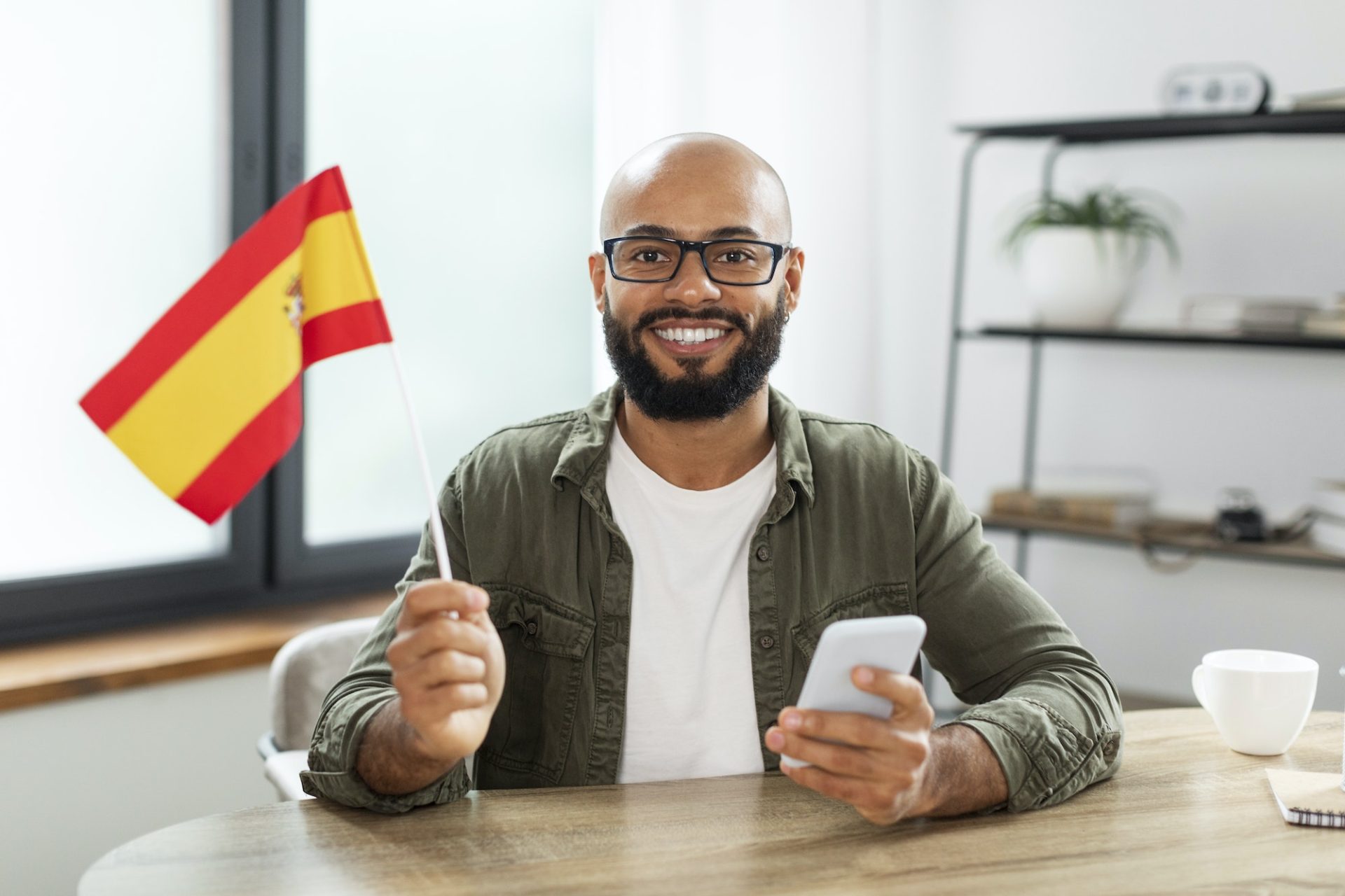Excited male tutor sitting at table with flag of Spain and using smartphone, looking and smiling at