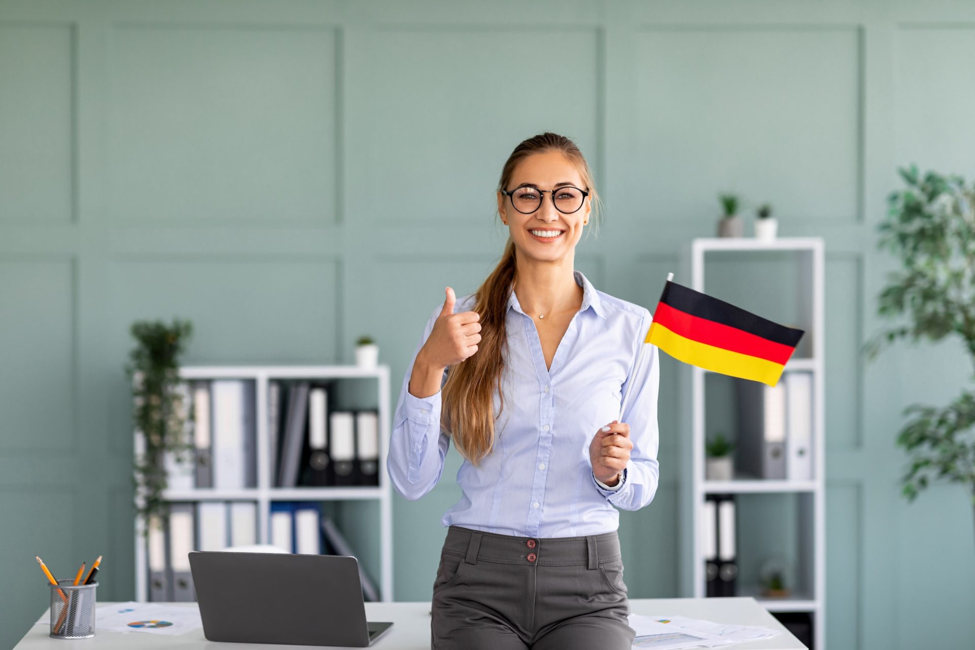 Happy female tutor with flag of Germany showing thumb up, standing near workplace in office interior
