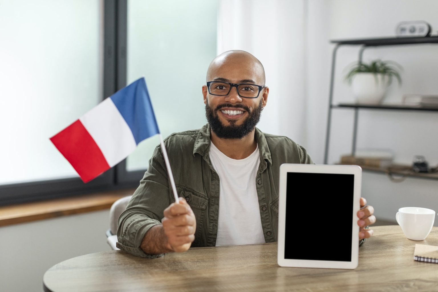 Happy latin man holding flag of France and showing tablet with blank screen, sitting at desk in room